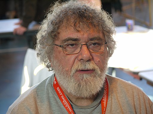 Valokuva rintakuvarajauksessa harmaapaitaisesta, harmaapartaisesta miehestä, jolla on korville asti ulottuvat kiharat harmaat hiukset ja mustat kulmakarvat. Hänellä on silmälasit ja kaulassa oranssi avainnauha. Hän katsoo kameran ohi, ilme on hyvin neutraali. Ympäristöstä ei saa selvää. Miehellä on luomi keskellä vasenta poskea, oikeassa poskessa suun vieressä sekä oikealla ohimolla.