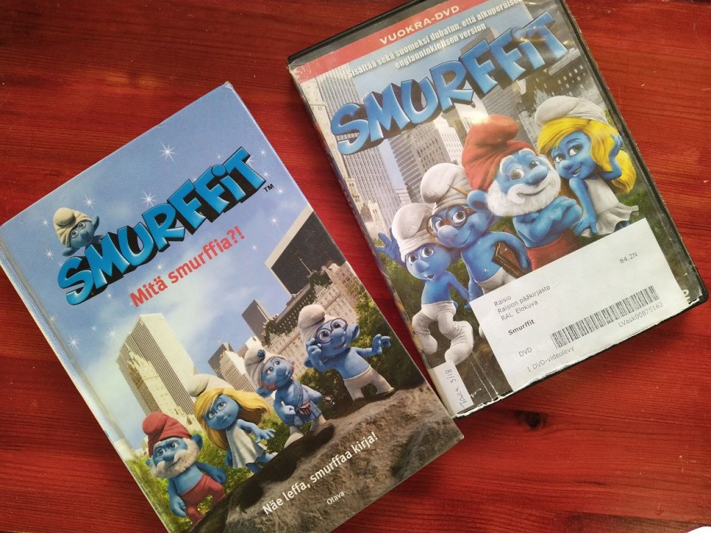 Valokuva DVD-elokuvan kannesta ja kirjan kannesta vierekkäin. Elokuvan nimi on Smurffit ja sen kannessa poseeraa neljä tietokoneanimoitua smurffihahmoa. Kirjan nimi on Smurffit Mitä smurffia?! ja sen alareunassa lukee Näe leffa, smurffaa kirja. Kirjan kanssa seisoo neljä tietokoneanimoitua smurffihahmoa puskaisella kivenlohkareella, taustalla aidon näköinen suurkaupunki pilvenpiirtäjineen.
