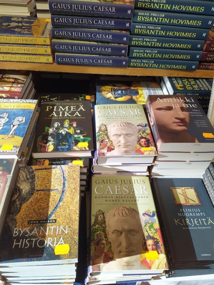 Valokuva kirjaläjistä myyntipöydällä. Keskimmäiset kirjapinot ovat Gaius Julius Caesar -kirjaa, sen molemmin puolin on erinimisiä kirjoja, kuten Bysantin historia, Pimeä aika, Antiikki ja me, Plinius nuoremman kirjeitä, Bysantin hovimies ja Keskiajan hirviöt.