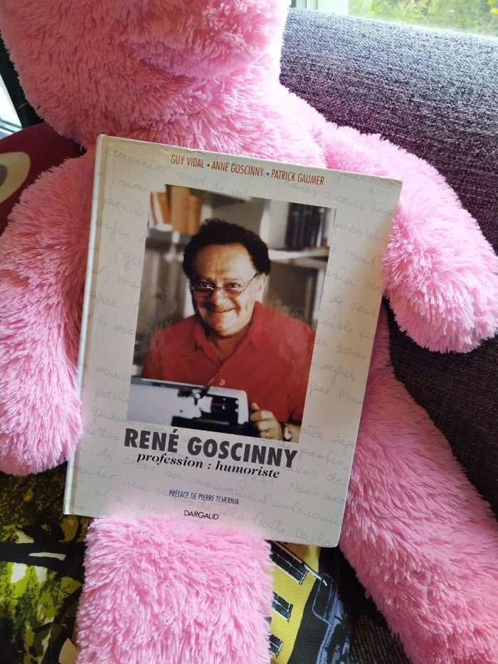 Valokuva kirjasta ison pinkin pehmoeläimen sylissä. Kirjan kannessa on kuva Rene Goscinnysta iloisena punaisessa paidassa kirjoituskoneen takana. Kirjan nimi on René Goscinny profession : humoriste, kirjoittaneet Guy Vidal, Anne Goscinny ja Patrick Gaumer, kustantanut Dargaud.