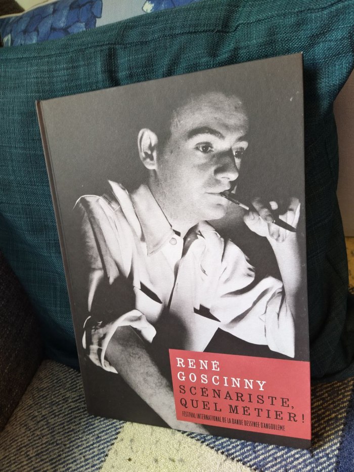 Valokuva kirjasta sohvatyynyä vasten. Mustavalkoisessa kansikuvassa nuori Goscinny pitelee kynää mietteliäästi huulillaan. Kirjan nimi on René Goscinny Scénariste, quel métier. Kannessa ei ole tekijätietoja, vain maininta Angoulemen sarjakuvafestivaaleista.